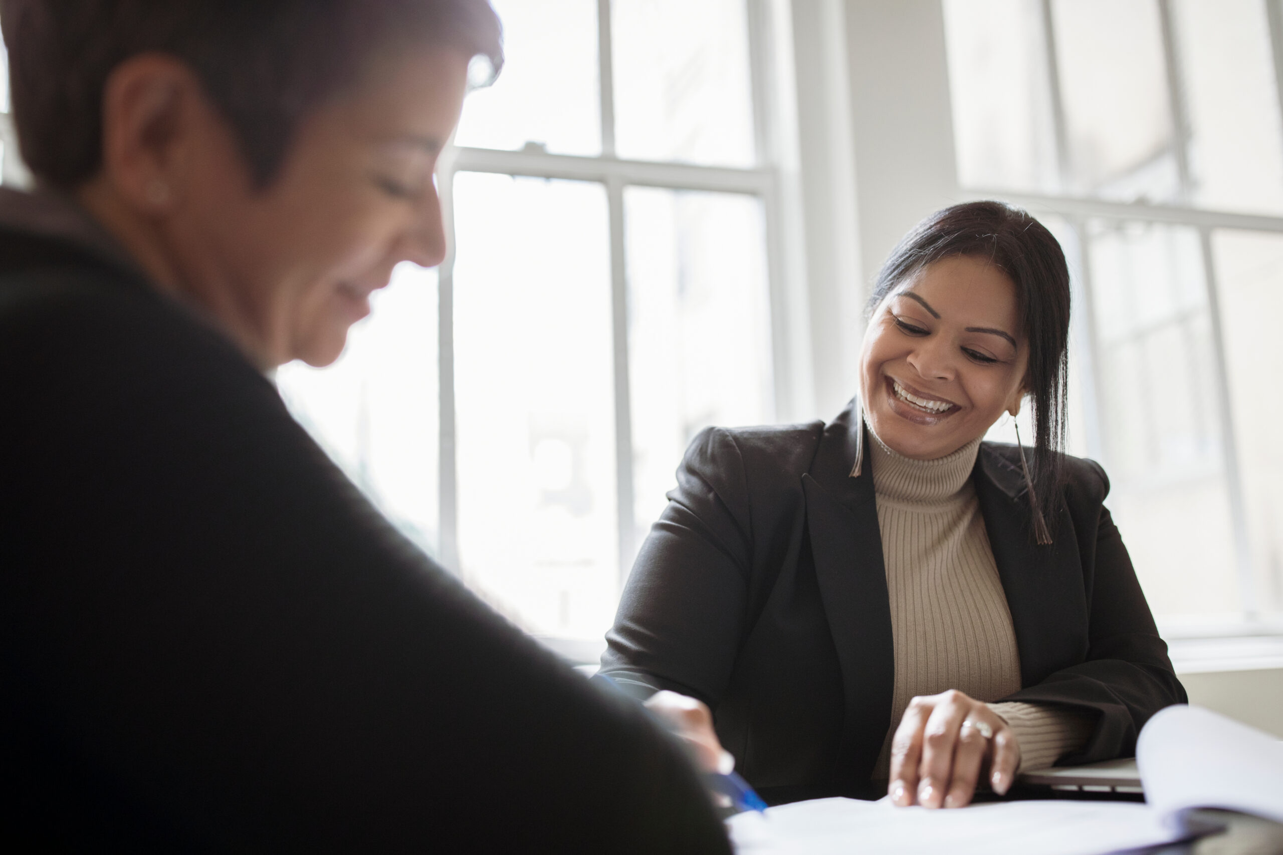 Low angle view of smiling female colleagues discussing documents while sitting against windows in office