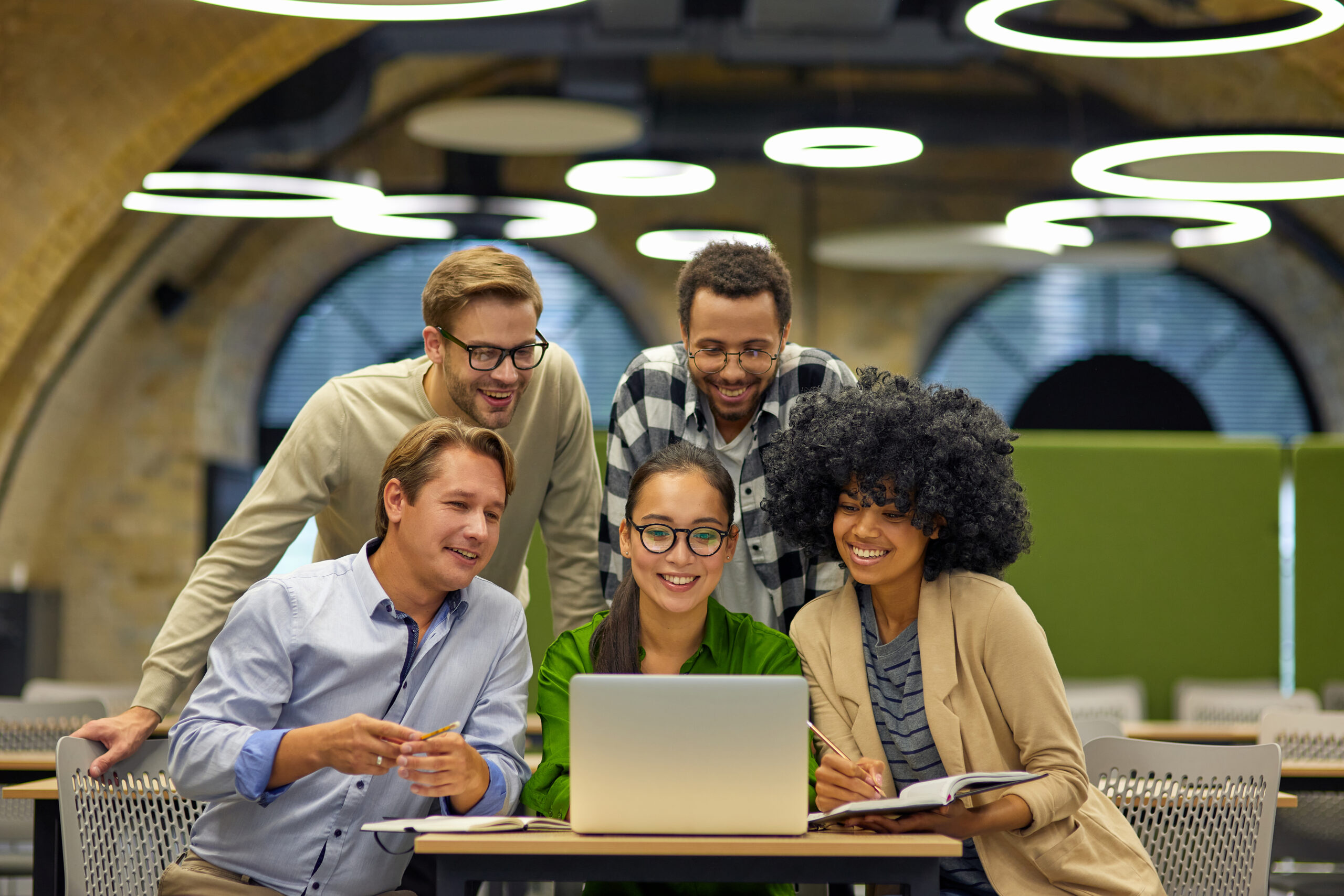 Group of young multiracial business people sitting at desk in the modern coworking space, looking at laptop screen and smiling, working together on a new project. Teamwork and collaboration