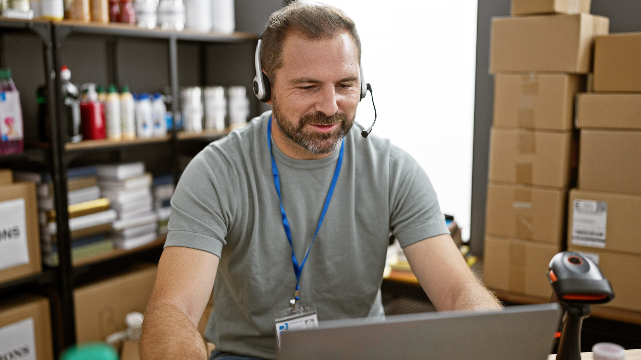 A handsome mature man with grey hair works in a warehouse, surrounded by donations and packages, sporting a headset and a badge.