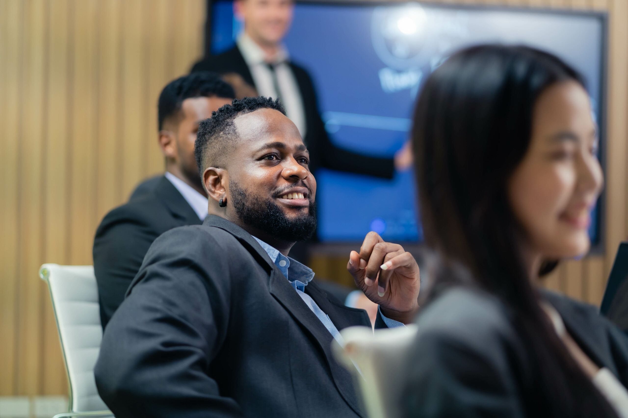 A man in a suit is smiling at a presentation. A woman is sitting next to him. The man is wearing a tie